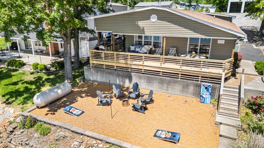 Aerial shot of a lakefront property with a long porch and a gravel yard with multiple outdoor chairs, a fire pit, and two cornhole boards