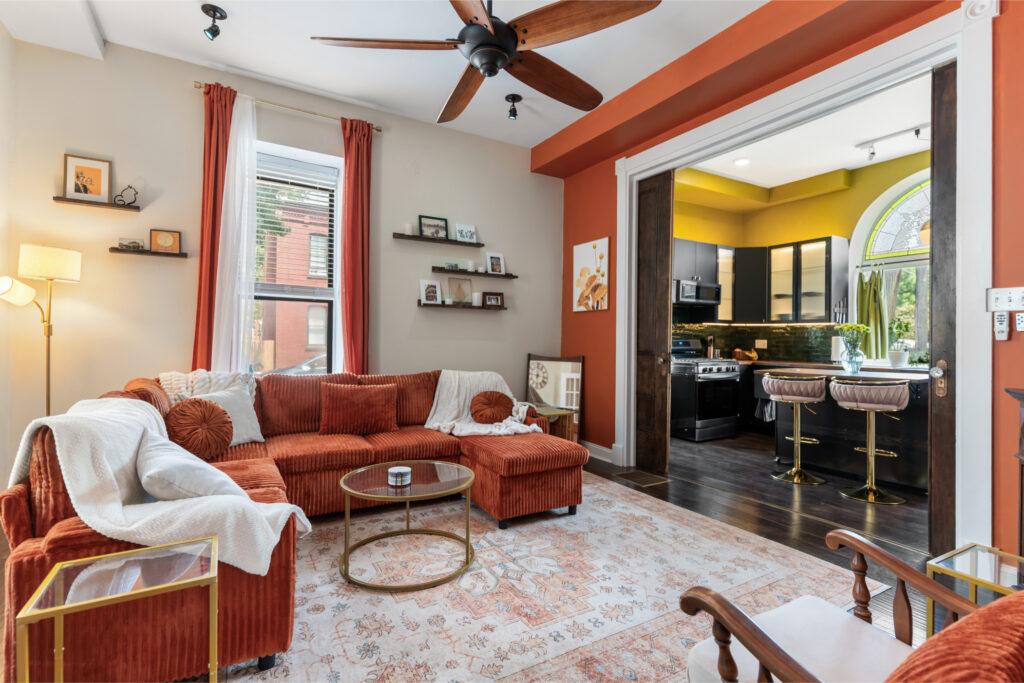 A living room with cream colored walls and orange furniture leading into a kitchen