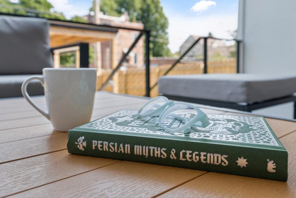 Close up of a book titled "Persian Myths and Legends" resting on a table with reading glasses on top and a coffee mug beside it